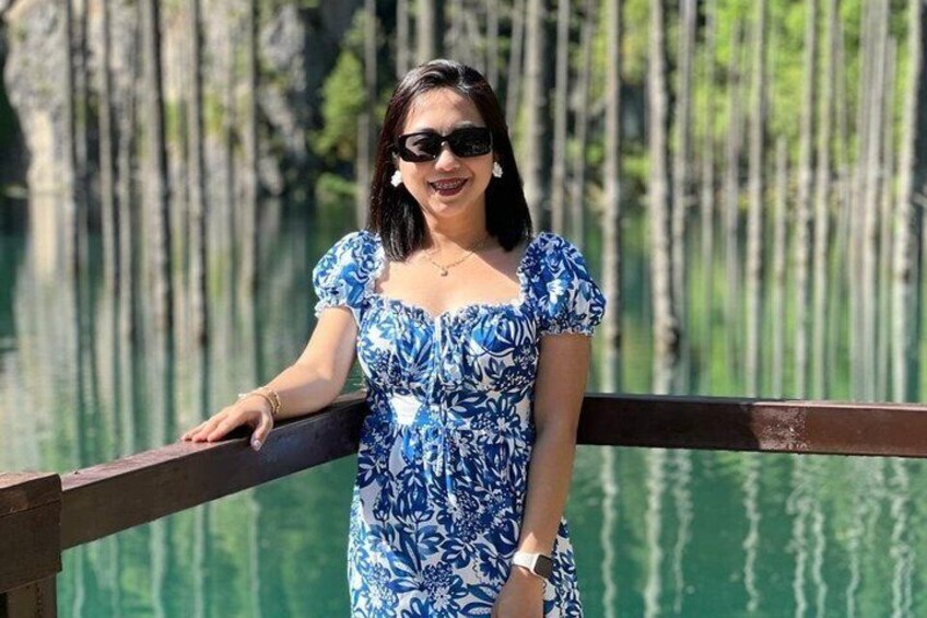 Eine Person in einem blauen Kleid mit Blumenmuster steht auf einer Holzbrücke, im Hintergrund befindet sich ein Gewässer und Bäume. 1