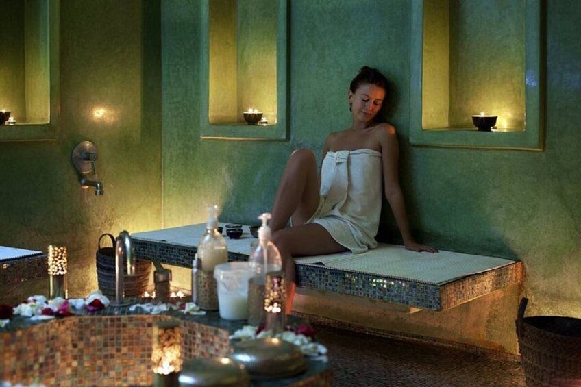 A person sitting on a bench in a spa-like setting with a tiled floor, a small table with bottles, and a wall-mounted shelf with a candle. 3