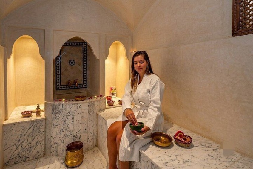 Une personne vêtue d’une robe blanche est assise sur un banc de marbre dans une pièce ornée d’arches et d’un mur décoratif. 4