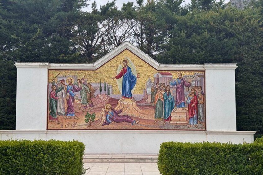 Een mozaïek met een centrale figuur in blauw, omringd door figuren in verschillende kleuren, geplaatst tegen een witte muur met groen op de achtergrond. 2