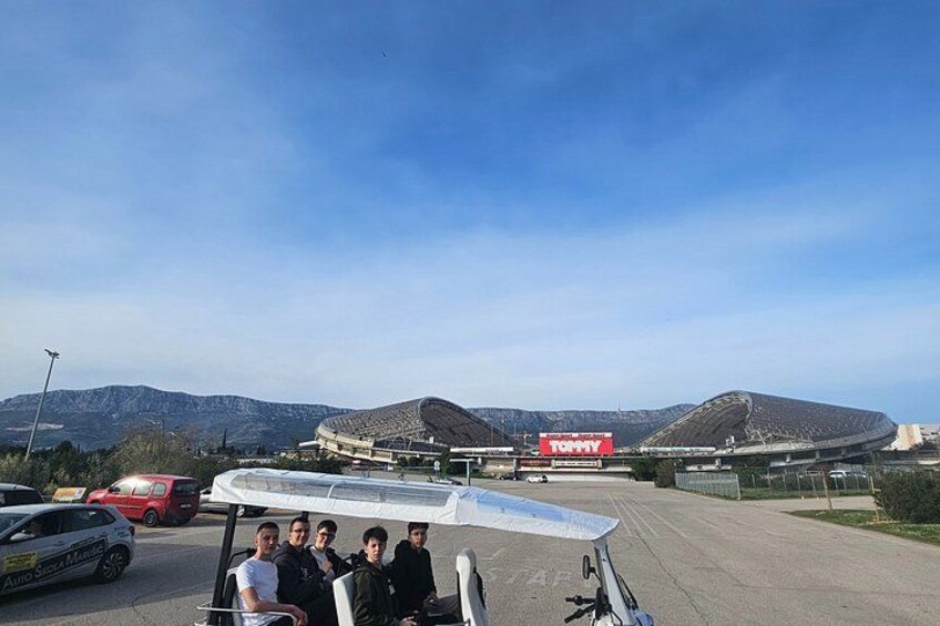 Un estadio con un gran letrero rojo, una carretera con coches aparcados y montañas al fondo. 3