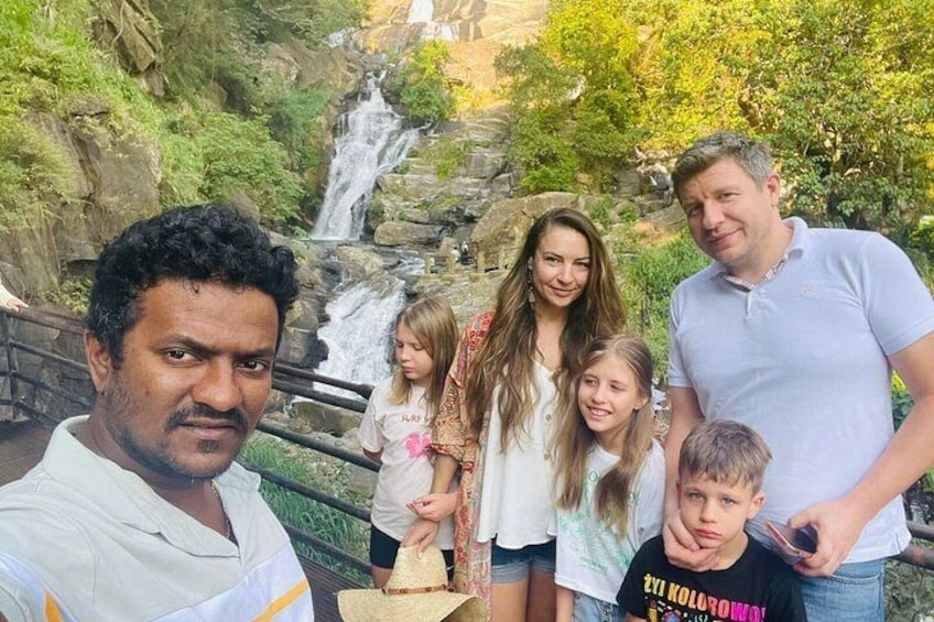 A family of five in front of a waterfall with lush greenery and a rocky landscape. 2