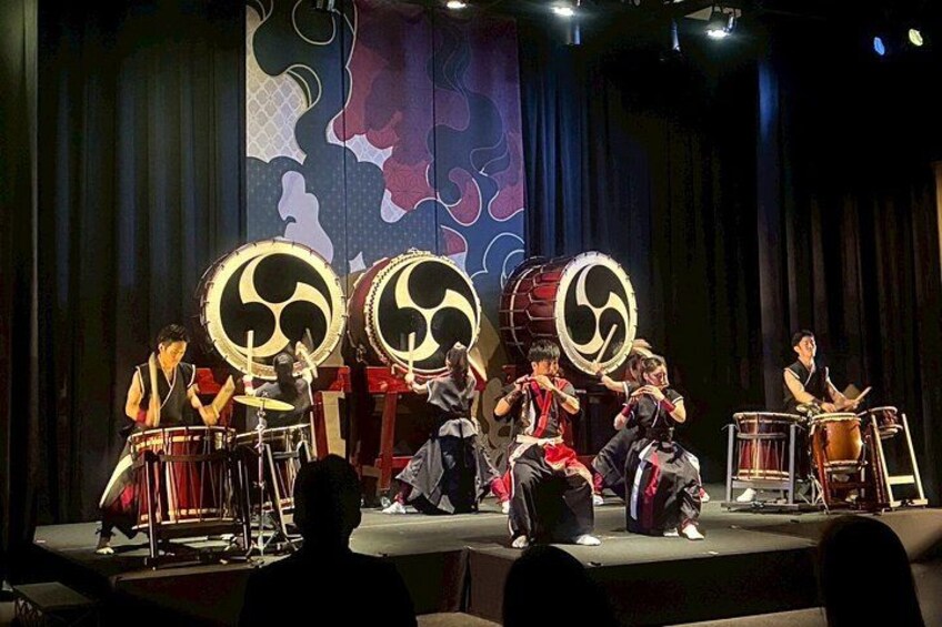 Un gruppo di musicisti si esibisce sul palco con tamburi tradizionali e uno sfondo con motivi astratti. 3