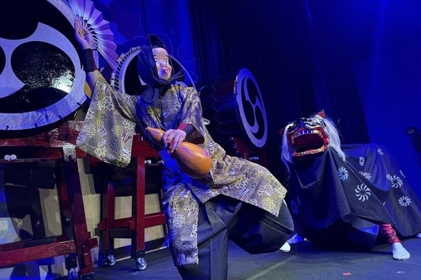 Un artista con traje tradicional y un abanico, otro con máscara, y un gran accesorio circular al fondo. 3