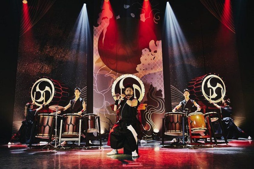 Presentación de percusión con tres percusionistas en el escenario, cada uno con un tambor que lleva un logotipo circular. 1