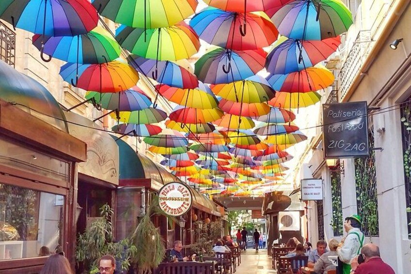 Una strada con ombrelloni colorati sospesi sopra, un caffè con tavolini all'aperto e un cartello per una pasticceria. 4