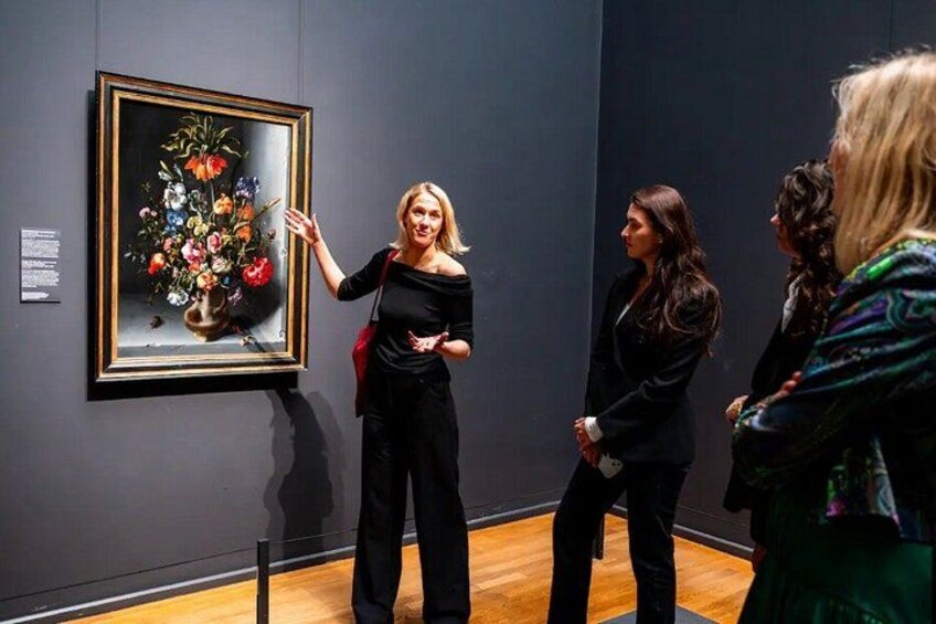 Iemand wijst naar een lijst met een bloemen schilderij aan de muur. 3