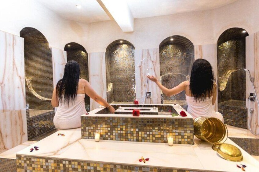 Two individuals in a spa-like setting with a mosaic-tiled wall, a marble floor, and a gold-colored basin. 3
