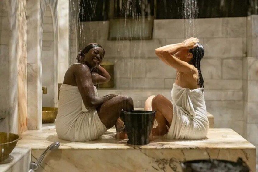 Two individuals sitting on a marble bench in a stone-walled room, with water flowing from above. 2