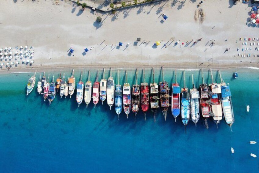 Vista aerea di una spiaggia con una fila di colorate imbarcazioni ormeggiate nell’acqua, una riva sabbiosa con ombrelloni e un mare azzurro e limpido. 4