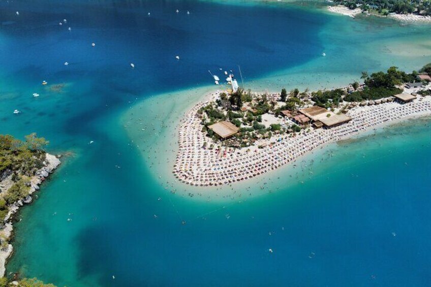 Vista aerea di una spiaggia con acque cristalline turchesi, una lingue di sabbia a forma di mezzaluna e un piccolo gruppo di edifici e ombrelloni. 3