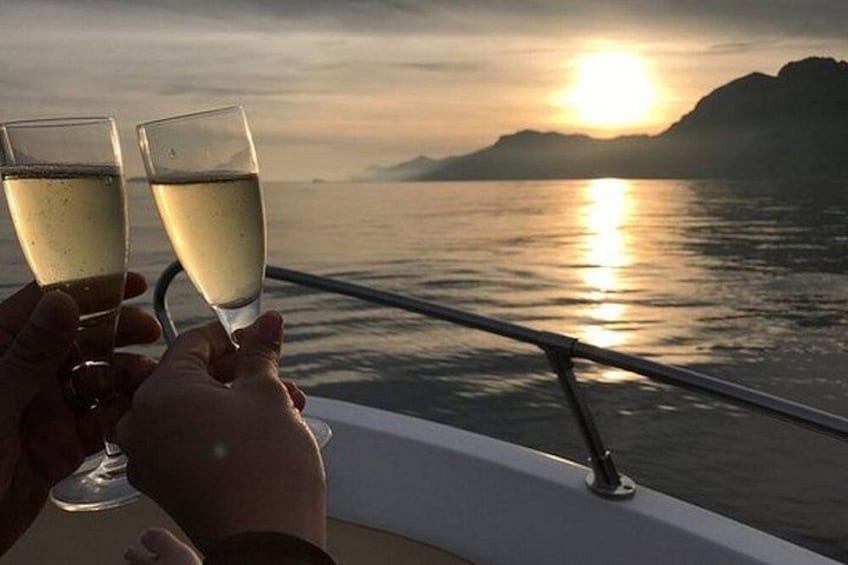To hænder holder champagneglas, en solnedgang over vandet, og et fjerntliggende bjerg. 4