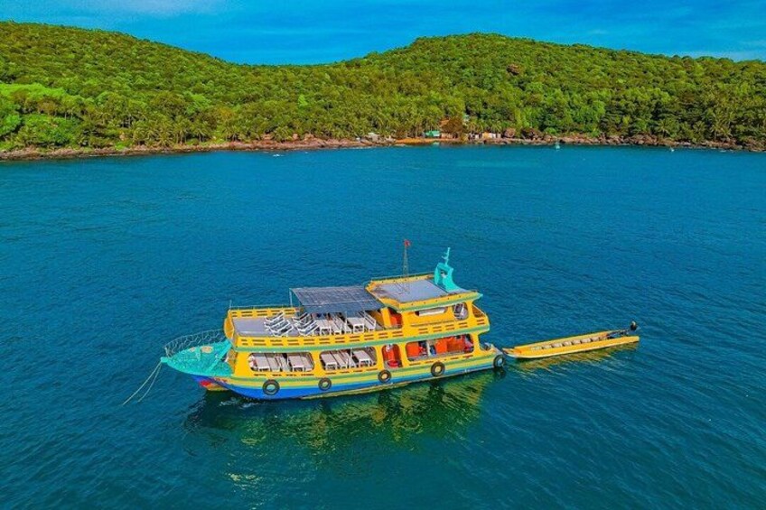 A multi-deck boat with a yellow and blue color scheme, floating on a calm body of water with a green, hilly shoreline in the background. 2