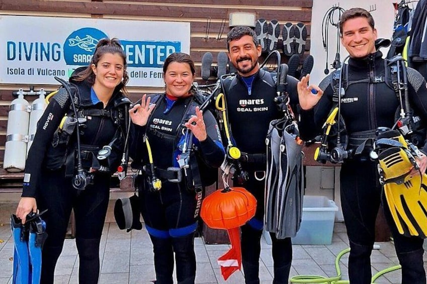 Buceadores con trajes de neopreno negros con logotipos 'mares' y 'seal skin', sosteniendo equipo de buceo, frente a un letrero de centro de buceo. 4