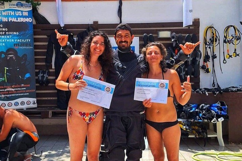Tres personas sosteniendo certificados, con equipo de buceo al fondo. 2