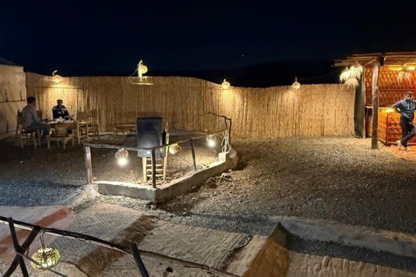 Un'ambientazione esterna rustica con una recinzione in legno, un tavolo con sedie e una piccola struttura illuminata. 4