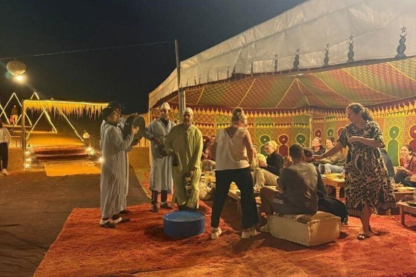 Un gruppo di persone riunite attorno a un tavolo sotto una tenda con uno sfondo colorato e decorato. 5