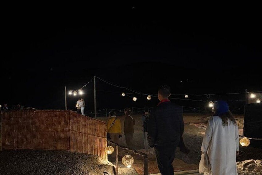 Spazio esterno illuminato di notte, con struttura in legno, luci a catena e persone che passeggiano. 3