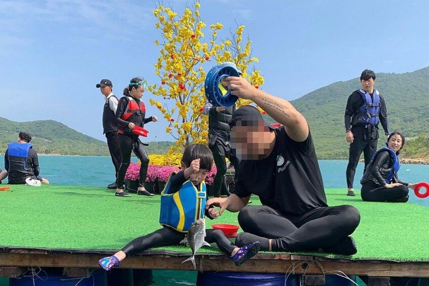 People on a green platform by a body of water, some holding blue and red objects, with yellow flowers and hills in the background. 2