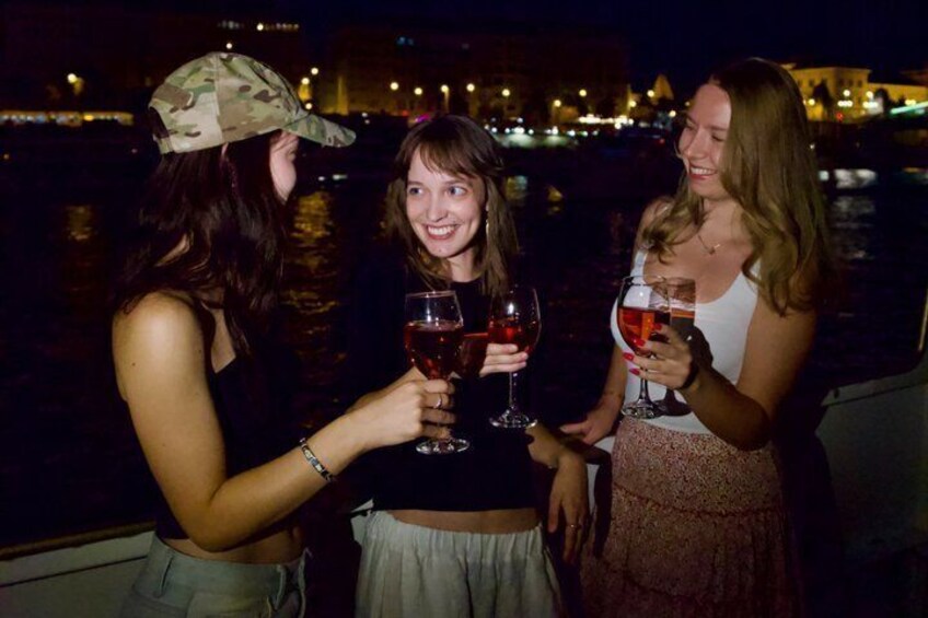Drie personen proosten met wijnglazen aan een waterkant in de nacht. 1