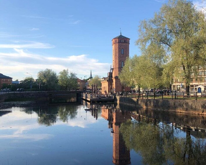 A historic tower by a calm river with a bridge and trees in the background. 1
