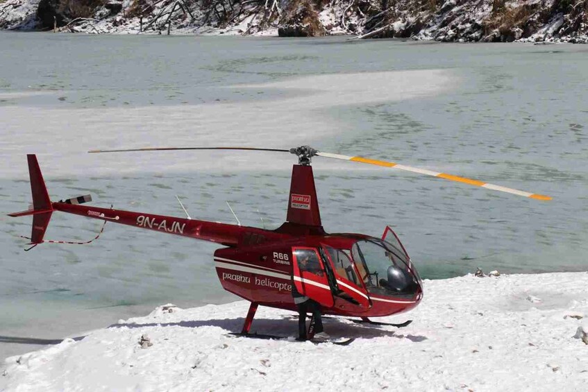 En röd helikopter med texten ”prahu helicopter” och ”9N-AJN” på sidan stod parkerad på en snötäckt yta nära ett vattendrag. 1