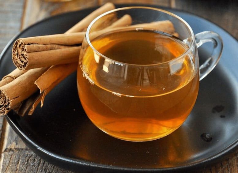 A clear glass cup of amber liquid, possibly tea, on a dark plate with cinnamon sticks beside it. 1