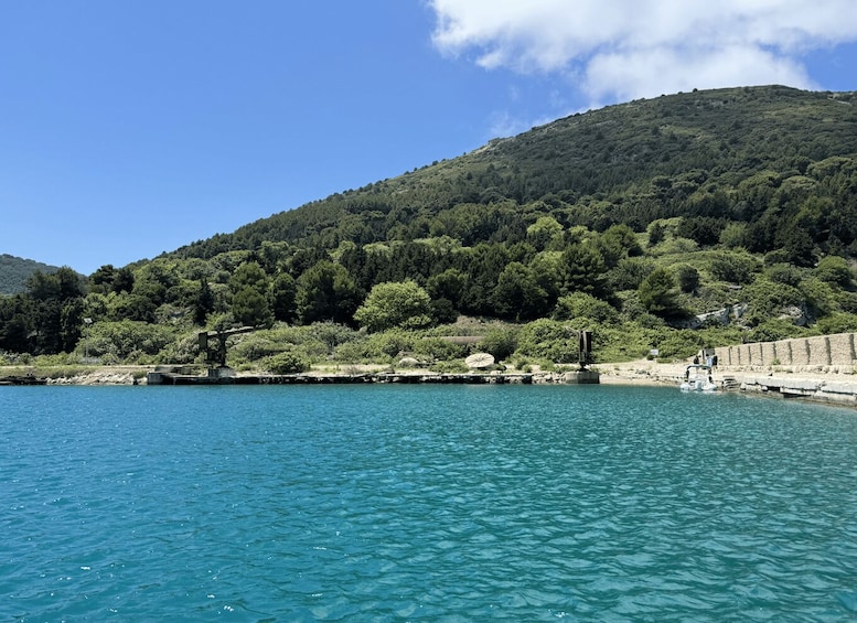 A clear blue lake with a rocky shoreline, surrounded by dense green forest and a hill in the background. 1