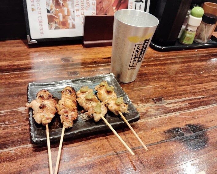A wooden table with a black plate holding skewered food, a metal cup, and some condiments. 5
