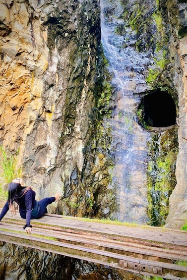 A person on a wooden platform near a waterfall and a cave entrance. 1