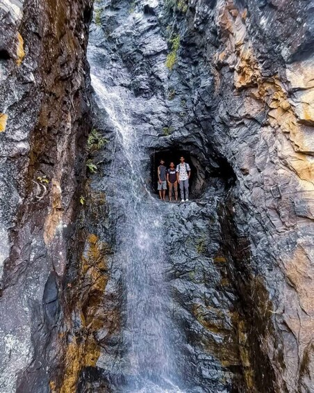 A waterfall in a rocky gorge with three people standing in a cave-like opening. 1