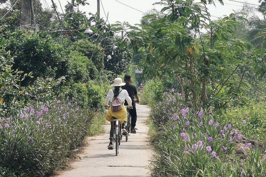 A pathway flanked by purple flowers and greenery, with two people riding bicycles. 1