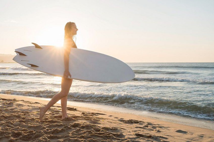 En person bærer en surfbræt på en strand med bølger og sol i baggrunden. 2