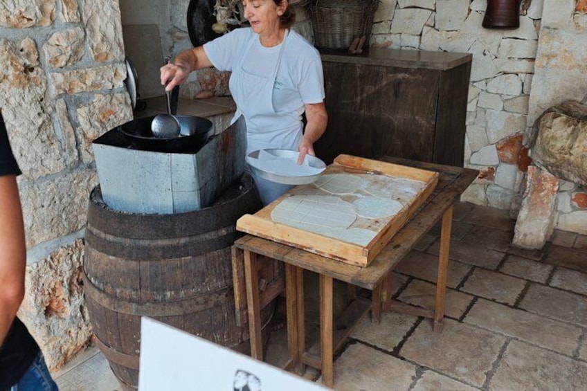 Una persona cocina al aire libre usando una olla metálica grande sobre un fogón, con una mesa de madera cerca. 3