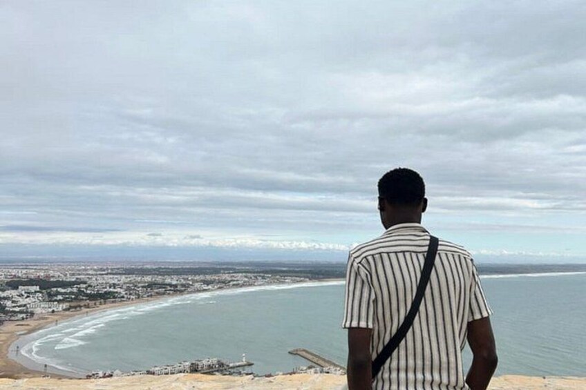 A man standing on a cliff overlooking a coastal city with a beach and a distant shoreline. 3