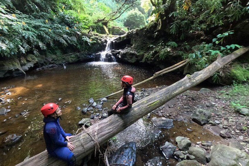 Two people in helmets sitting on a fallen log over a stream in a forest. 5
