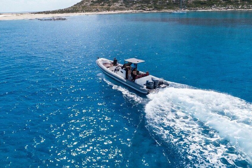 A speedboat on a clear blue sea with a wake behind it, a sandy beach, and a hilly landscape in the distance. 1