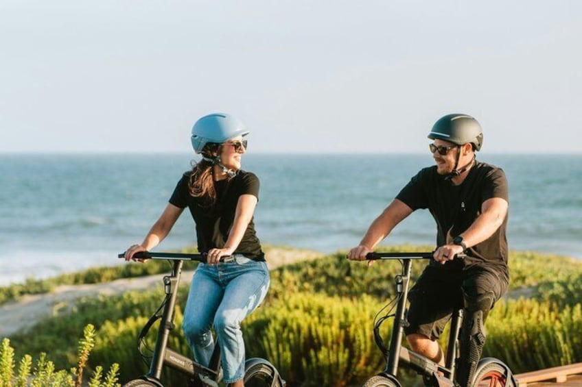 Deux personnes font du vélo électrique sur une piste cyclable en bord de mer, avec l’océan en arrière-plan. 4