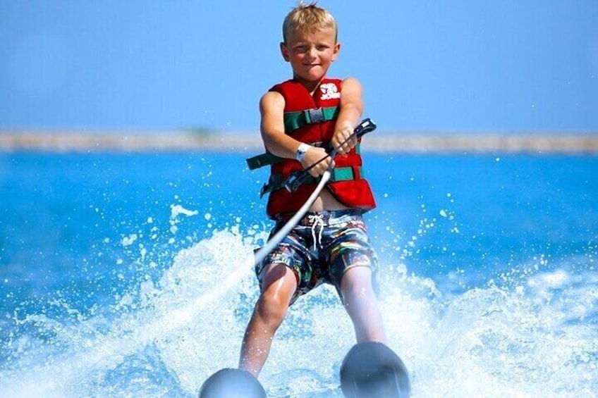 Een kind op waterski, met een reddingsvest en een ski-stok in de hand, omringd door opspattend water. 5