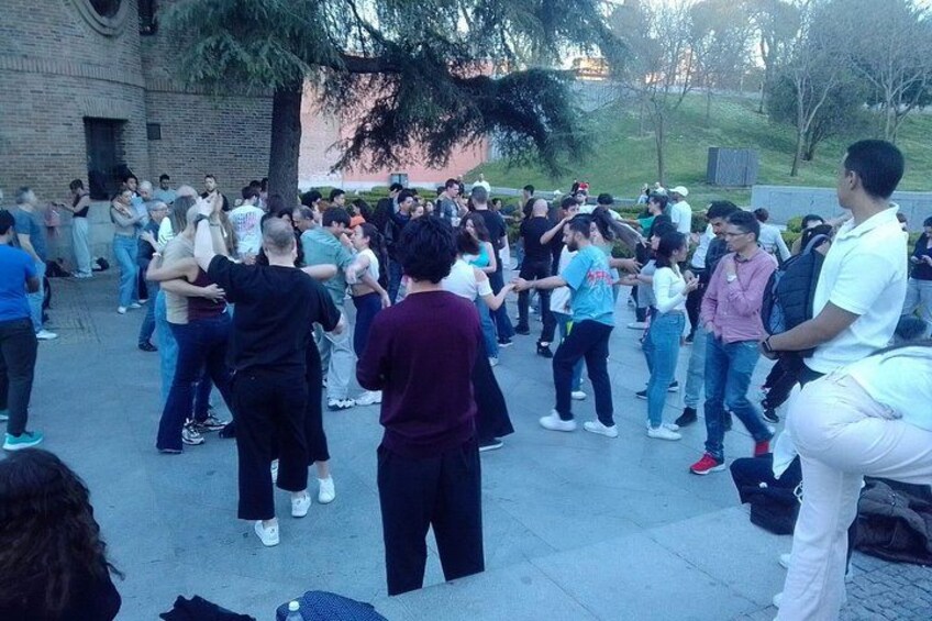A group of people performing a coordinated dance in an outdoor setting with a brick building and trees in the background. 1