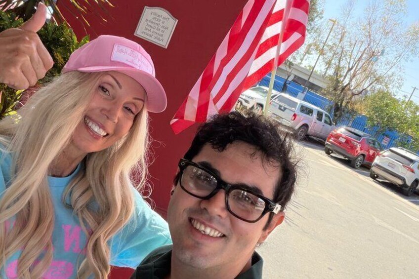 Two people in front of a red building with an American flag, a parking lot, and a blue fence in the background. 3