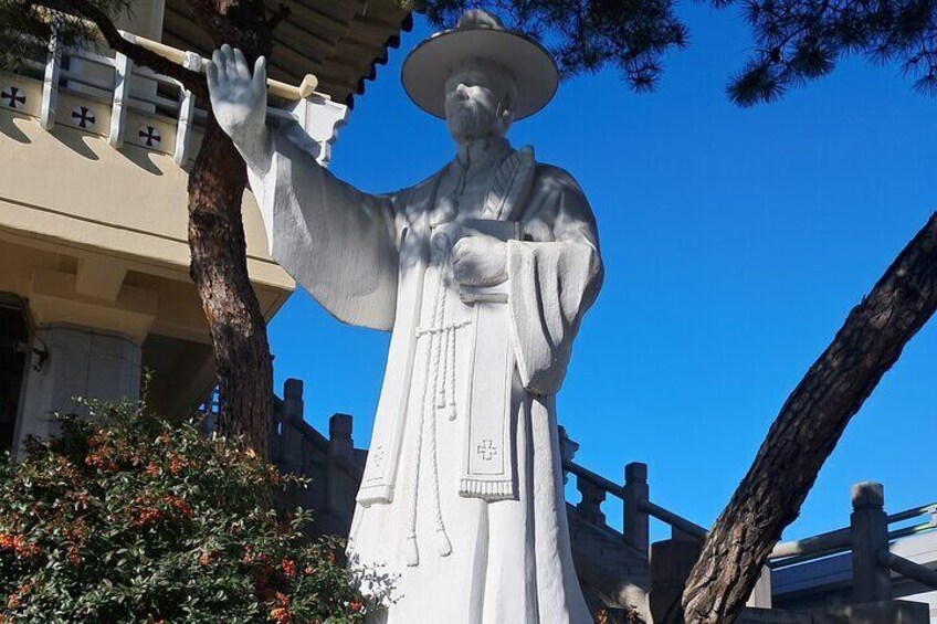 A statue of a figure in traditional attire with a hat, standing in front of a building with a fence and trees. 1