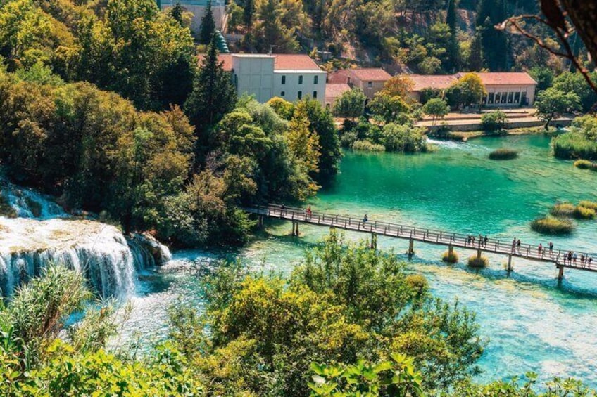 A waterfall, a bridge, and buildings by a river. 1