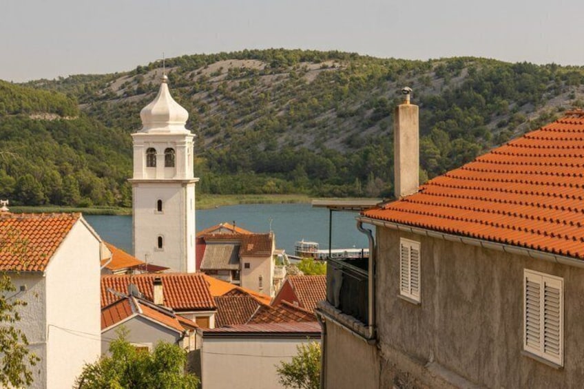A coastal town with terracotta-roofed buildings, a white church tower, and a backdrop of green hills and a calm sea. 3