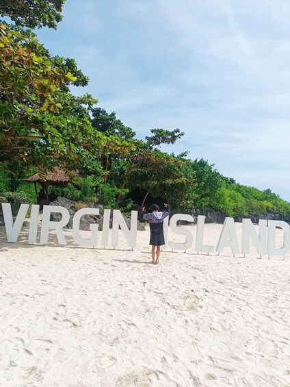 A person standing in front of large white letters spelling 'VIRGIN ISLAND' on a sandy beach with trees in the background. 2