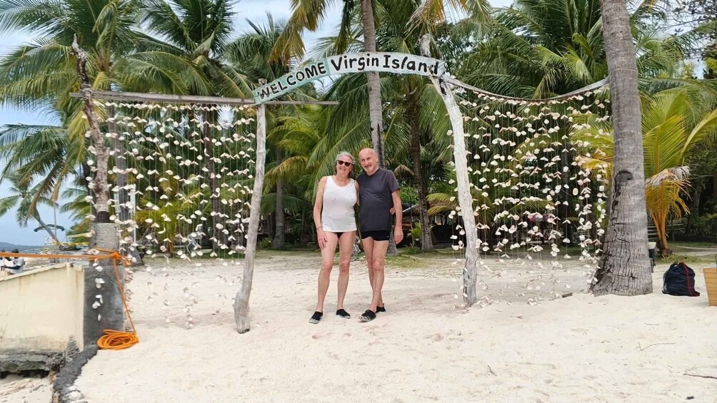 A couple standing under a 'Welcome Virgin Island' sign amidst a tropical setting with palm trees and hanging decorations. 3
