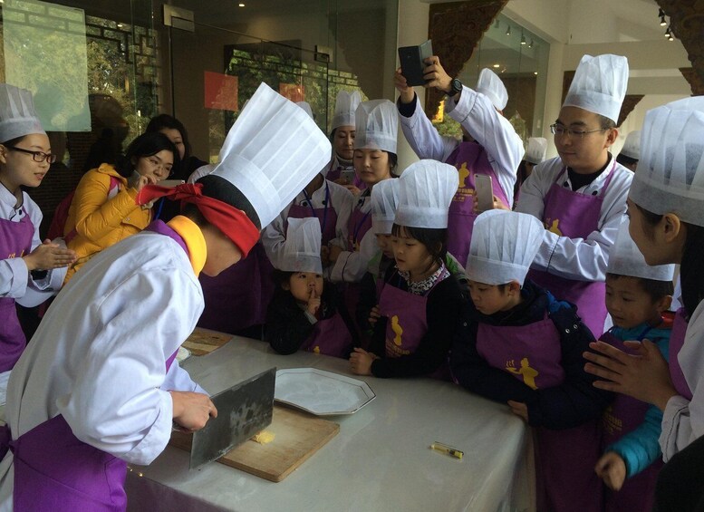 A group of people in chef hats and aprons gathered around a table with a chef demonstrating a cooking technique. 2