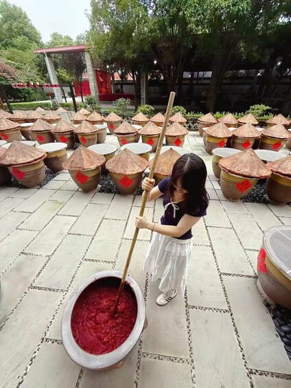 A person stirring a large bowl of red liquid outdoors, surrounded by traditional conical containers with red markings. 5