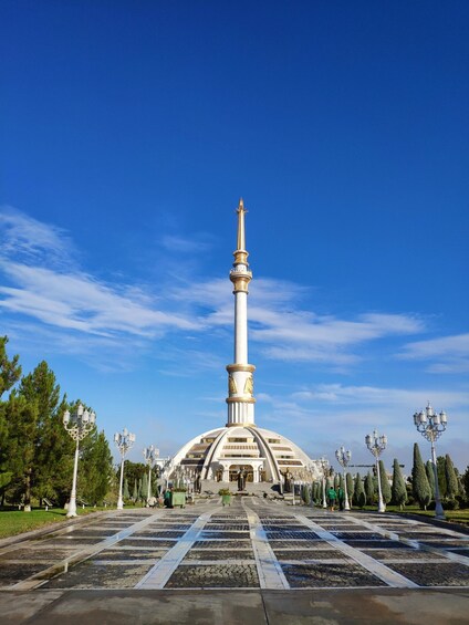 A tall white monument with a golden top, surrounded by a symmetrical garden with patterned paving and lamp posts. 3