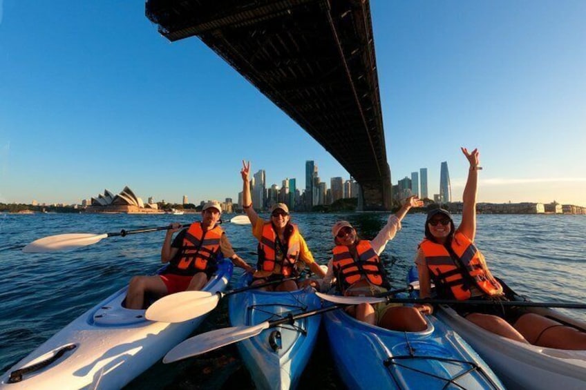 Three people kayaking under a bridge with a city skyline in the background. 3